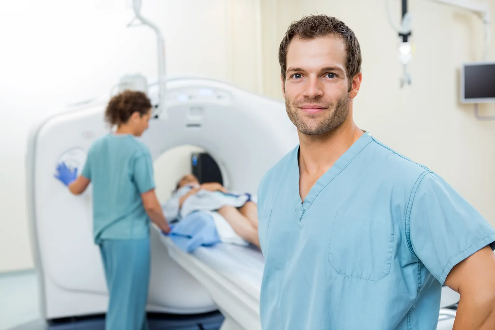 CT technologist next to a CT scanner, a colleague preparing a patient for the scan